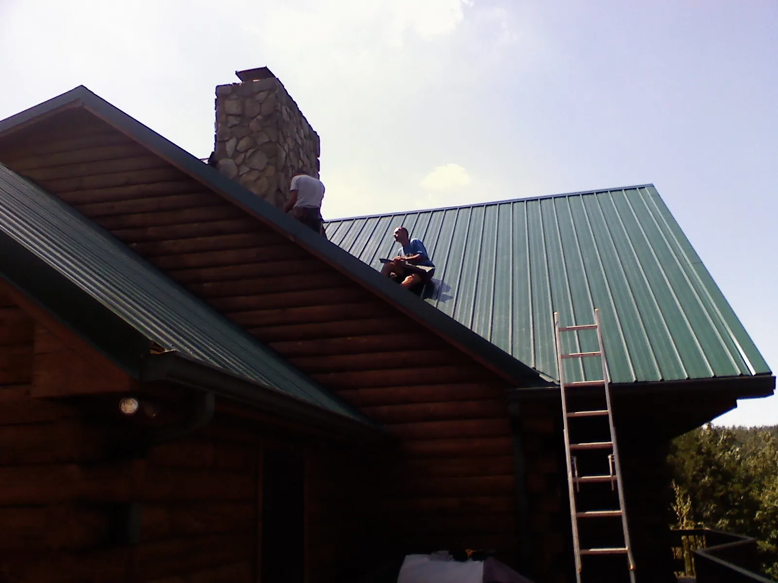 Roofers installing green metal panels beside a stone chimney on a log home in Adairsville